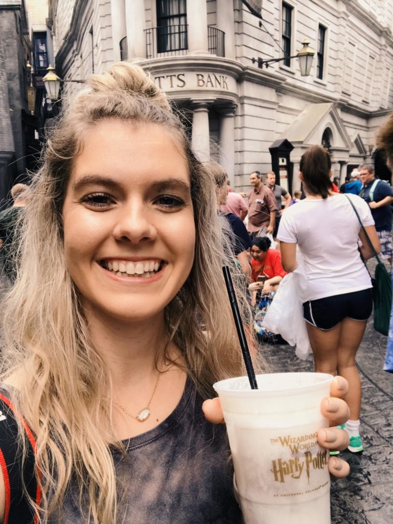 Selfie of blonde girl holding a butterbeer in front of Gringotts in Diagon Alley