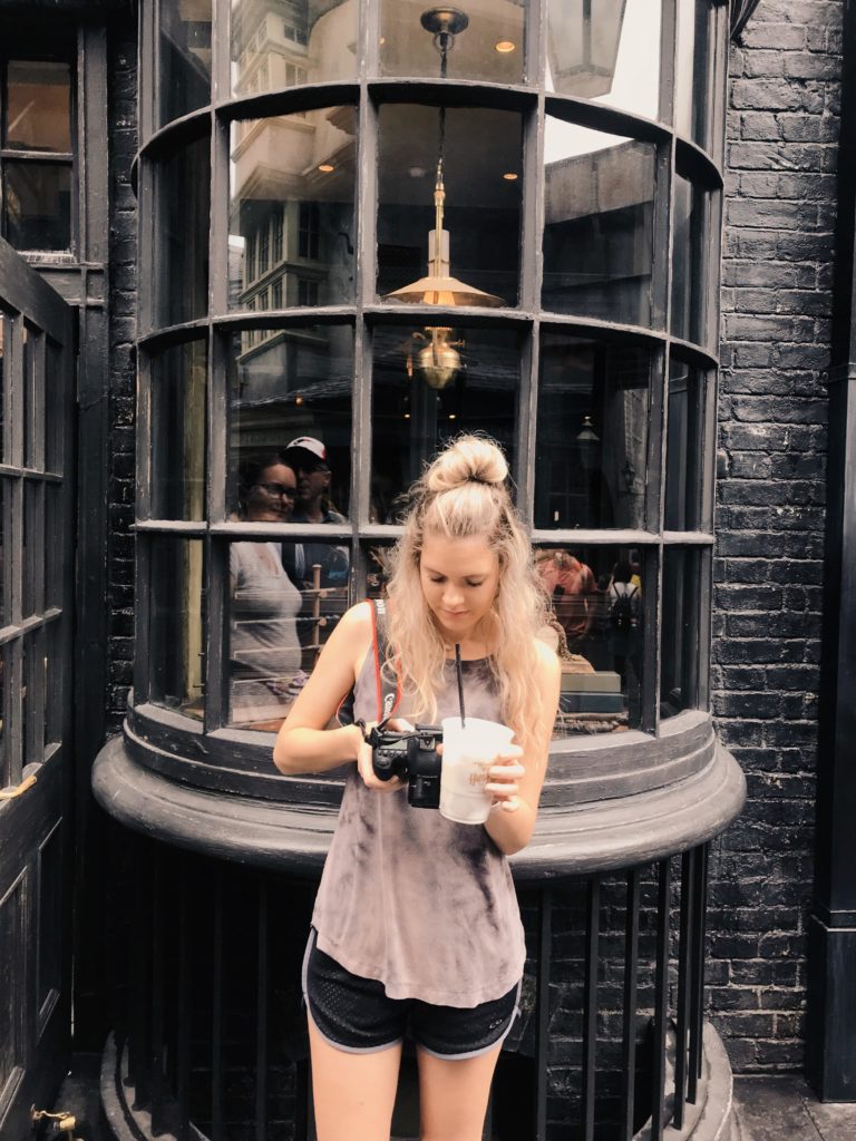 Girl looks at her camera while holding a butterbeer in front of Ollivander's