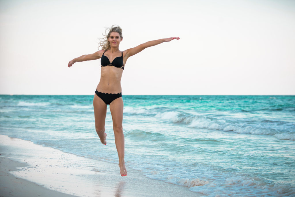 Blonde girl in a bikini leaping into the air in a dancer's pose with hair blowing in the wind