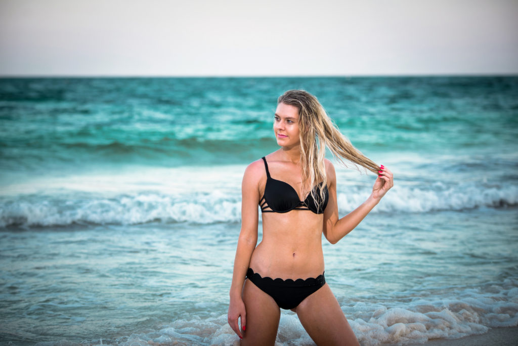 Blonde swimsuit model plays with her hair with the ocean in the background