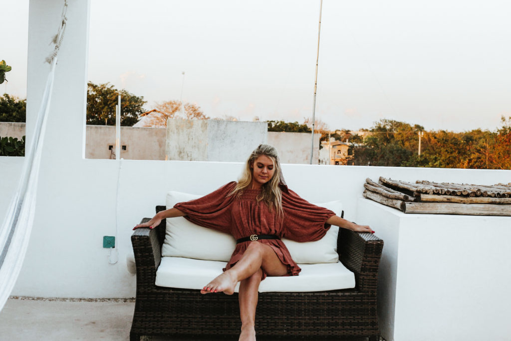Girl in a rust romper sits on a sofa on a rooftop with Tulum Town behind her