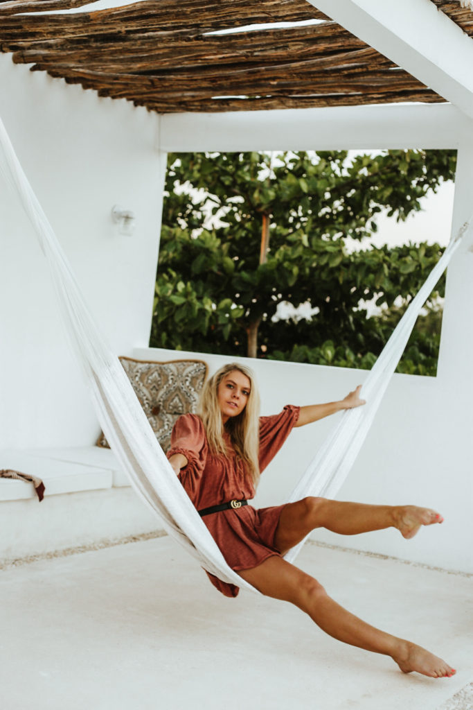 Model poses in a white hammock with a white limestone and wooden roof above her in the treetops