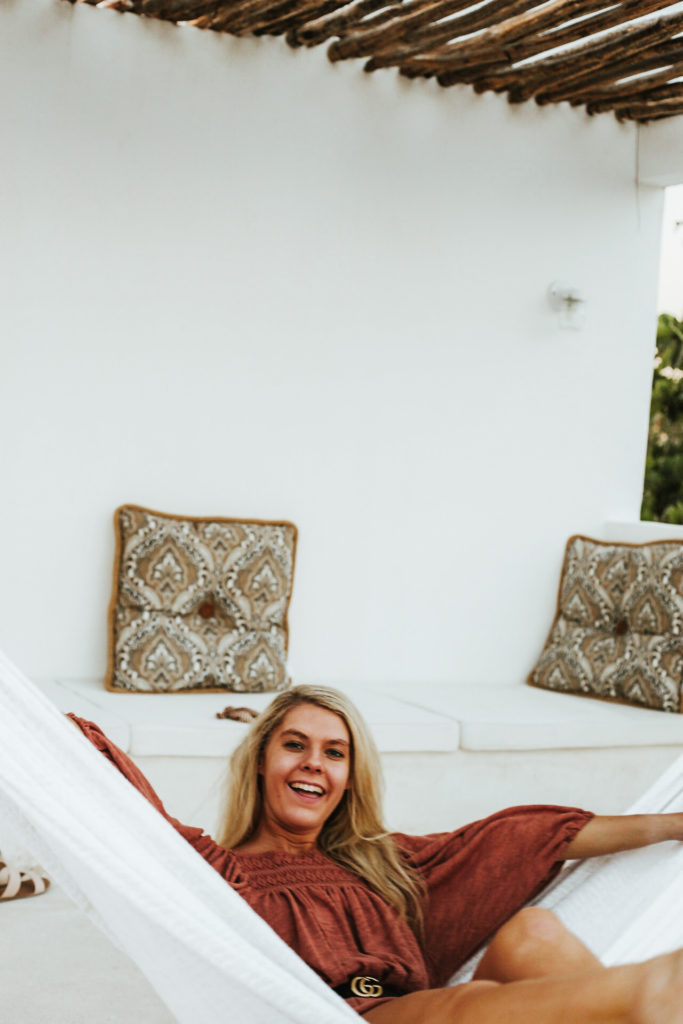 Blonde girl smiles while swinging in a hammock with paisley cushions and a patio seat behind her