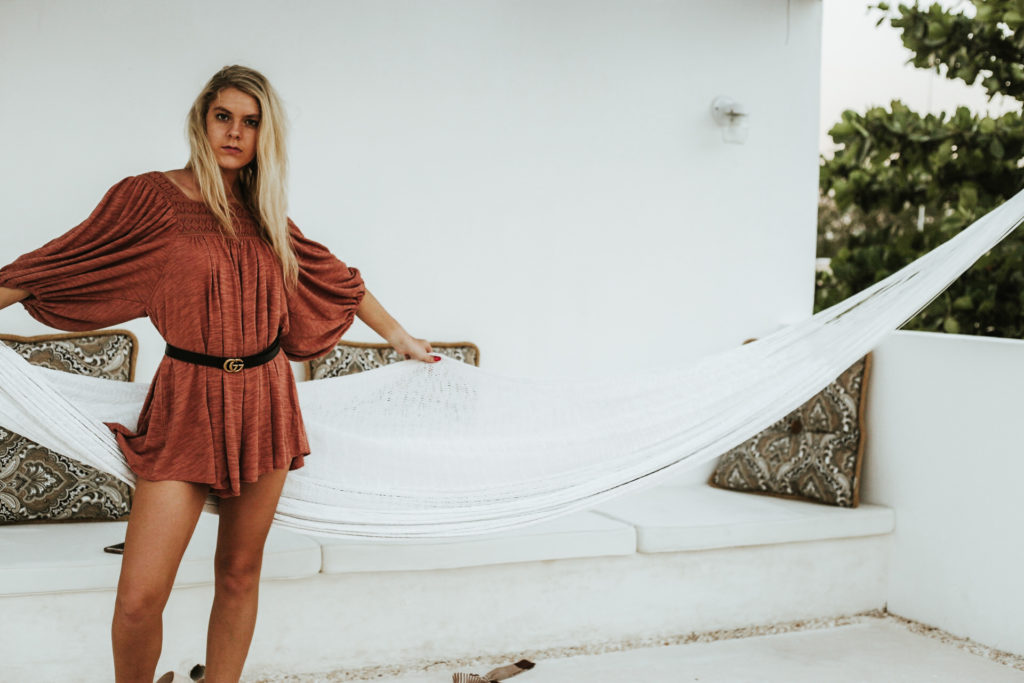 Model strikes a pose in front of a white hammock while wearing a bell sleeve belted romper
