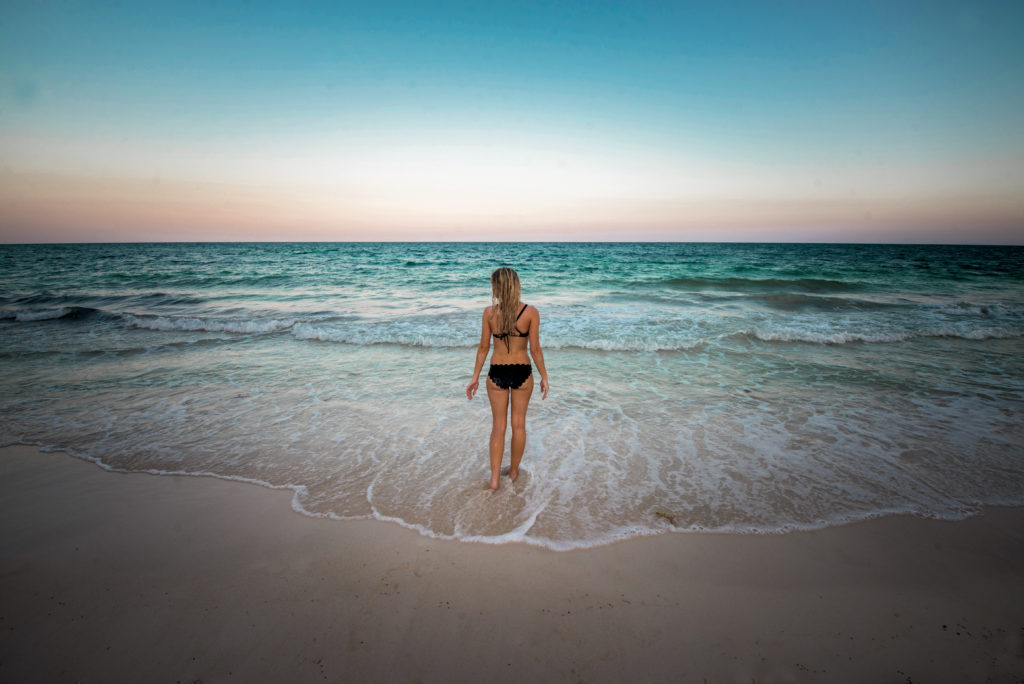 Blonde girl in a black bikini walks towards the sunset and crashing ocean waves in front of her